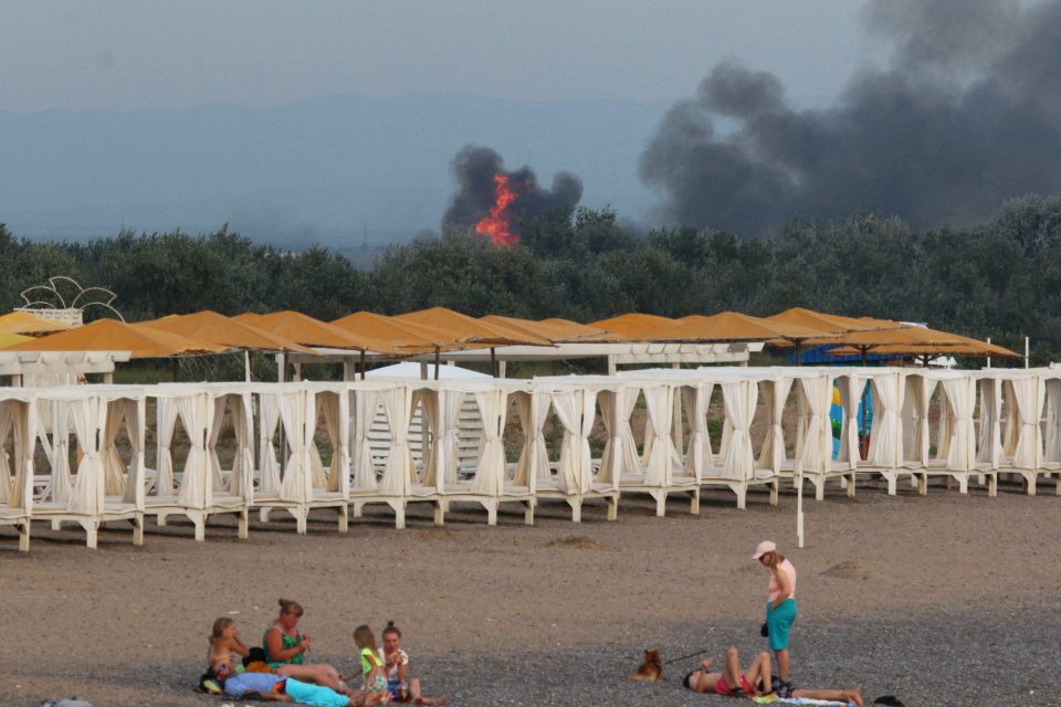 Lidé odpočívají na pláži,  zatímco po explozích na ruské vojenské letecké základně v Novofedorivce na Krymu stoupá kouř a jsou vidět plameny,  9. srpna 2022 | foto: Reuters